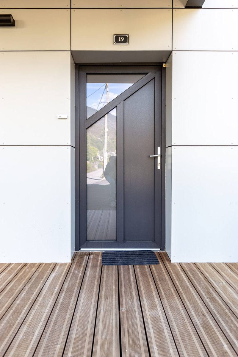 Dark front door with an angled glass panel on a white wall above a timber deck